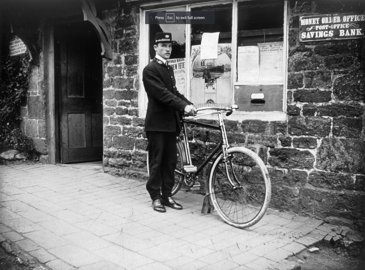 An old fashioned photo of a postman next to a post office with a bicycle.