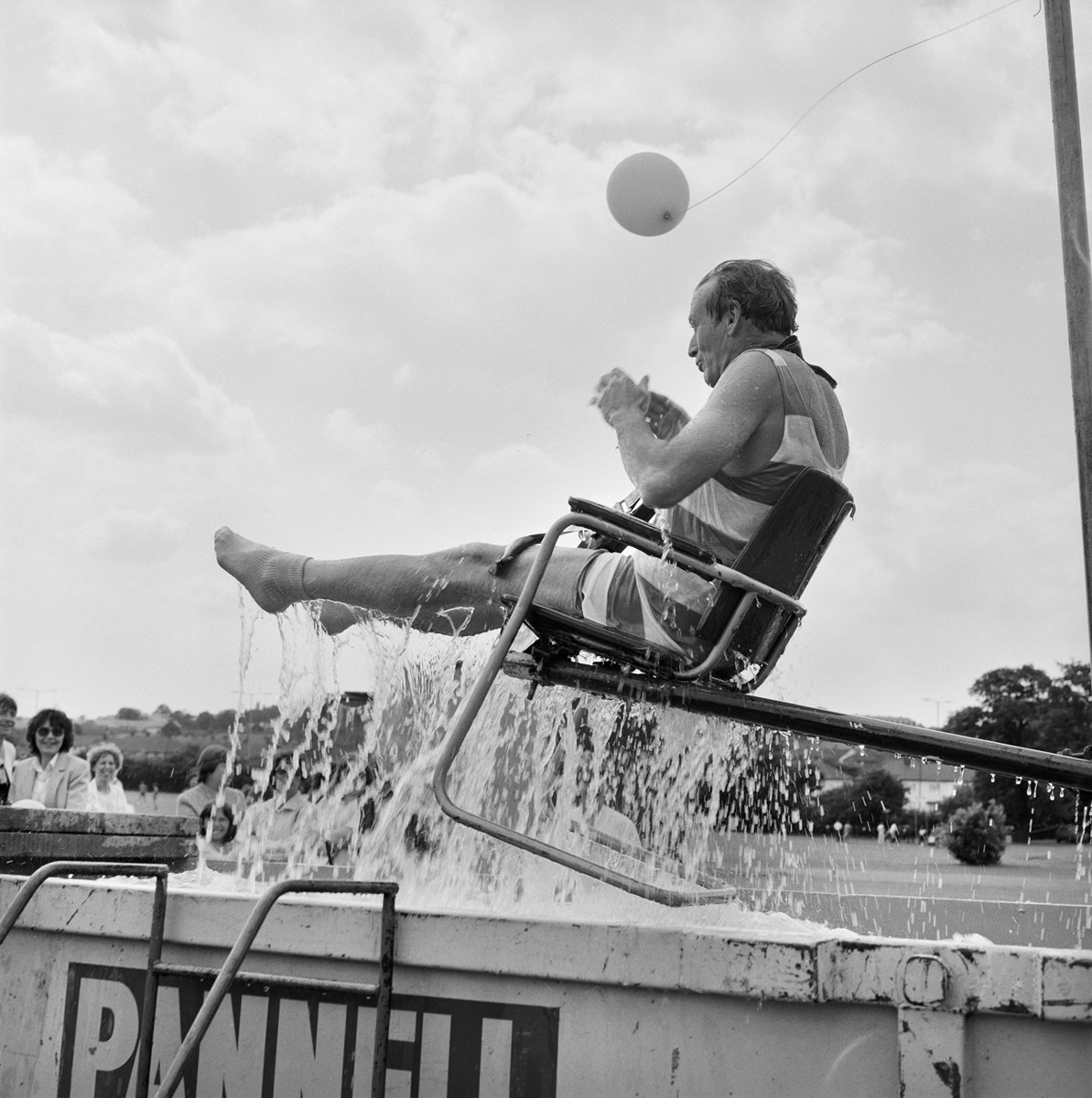 A man on a chair being lifted out of a skip filled with water.